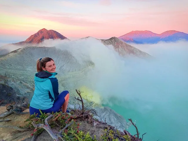 Woman sitting on a ridge overlooking a stunning turquoise crater lake with steam.