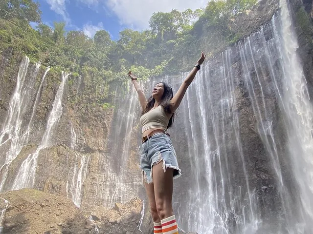 Woman standing with raised arms in front of a majestic waterfall cascading down rocks.