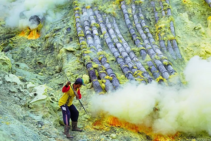 Miner collecting sulfur near pipes in a volcanic crater with thick yellow smoke.