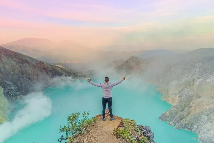 Hiker standing on a ridge with arms outstretched, looking at a turquoise crater lake.