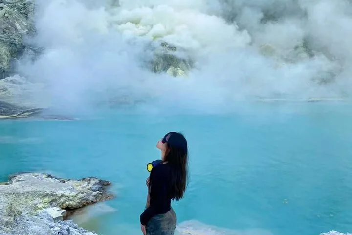 Person standing near a stunning turquoise crater lake surrounded by sulfurous steam.